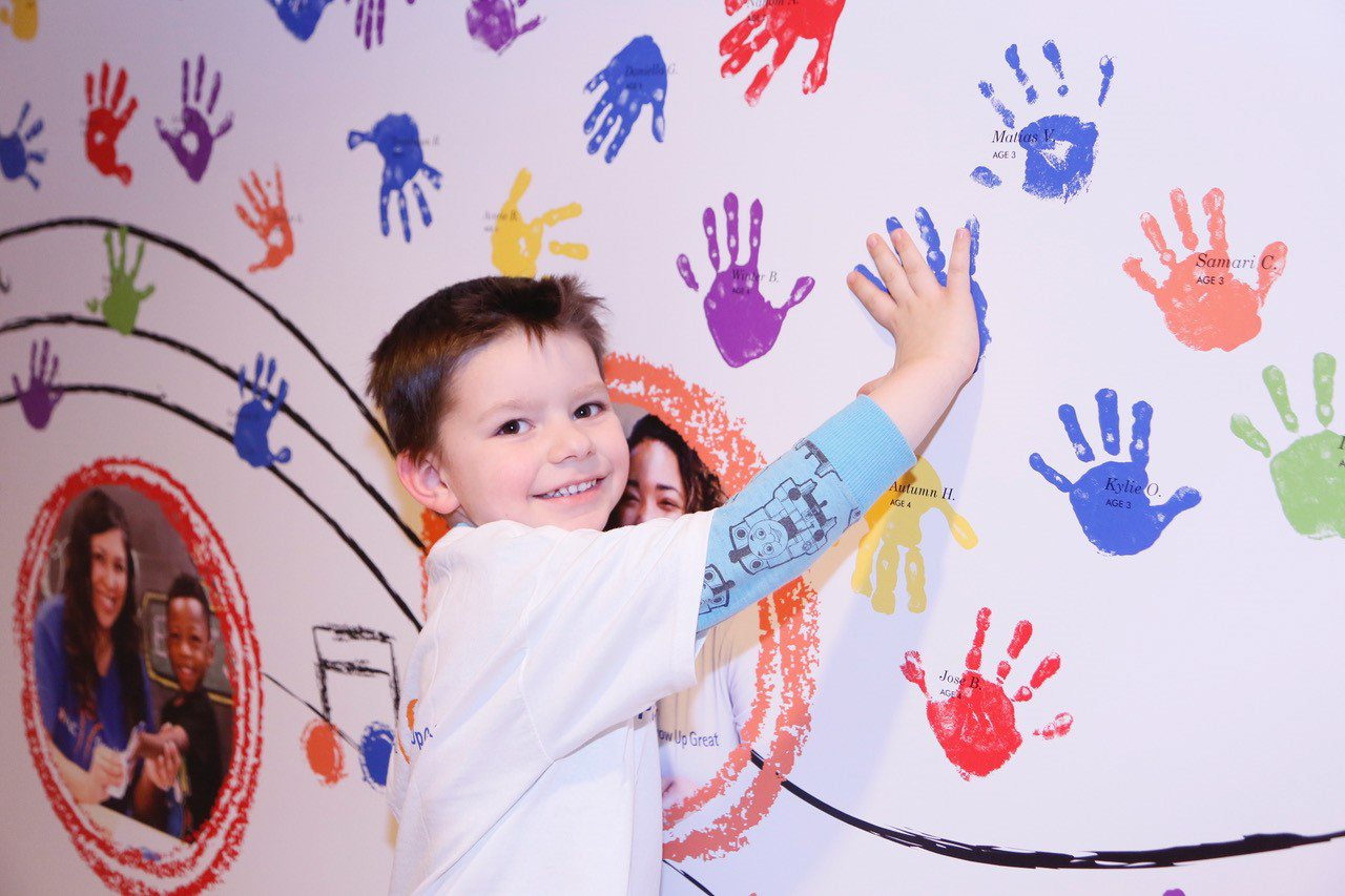 A young boy smiles as he places his hand on a wall covered with colorful handprints, adding his touch to the vibrant, community-created architecture and spaces. Other children and adults' handprints and photos are visible in the background.