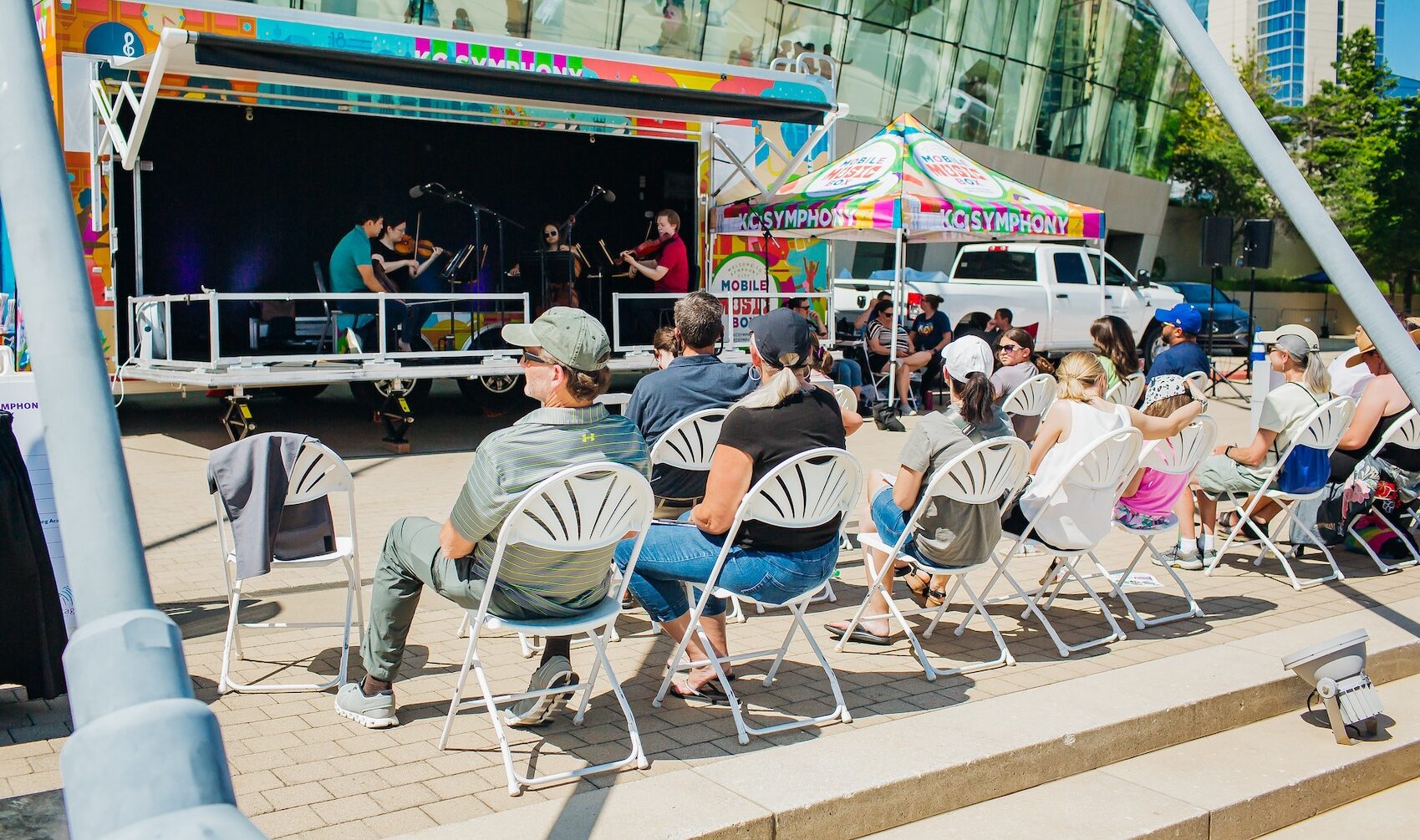 People sitting on white chairs outdoors, watching Kansas City Symphony musicians perform on the Mobile Music Box stage at Future Stages Festival.