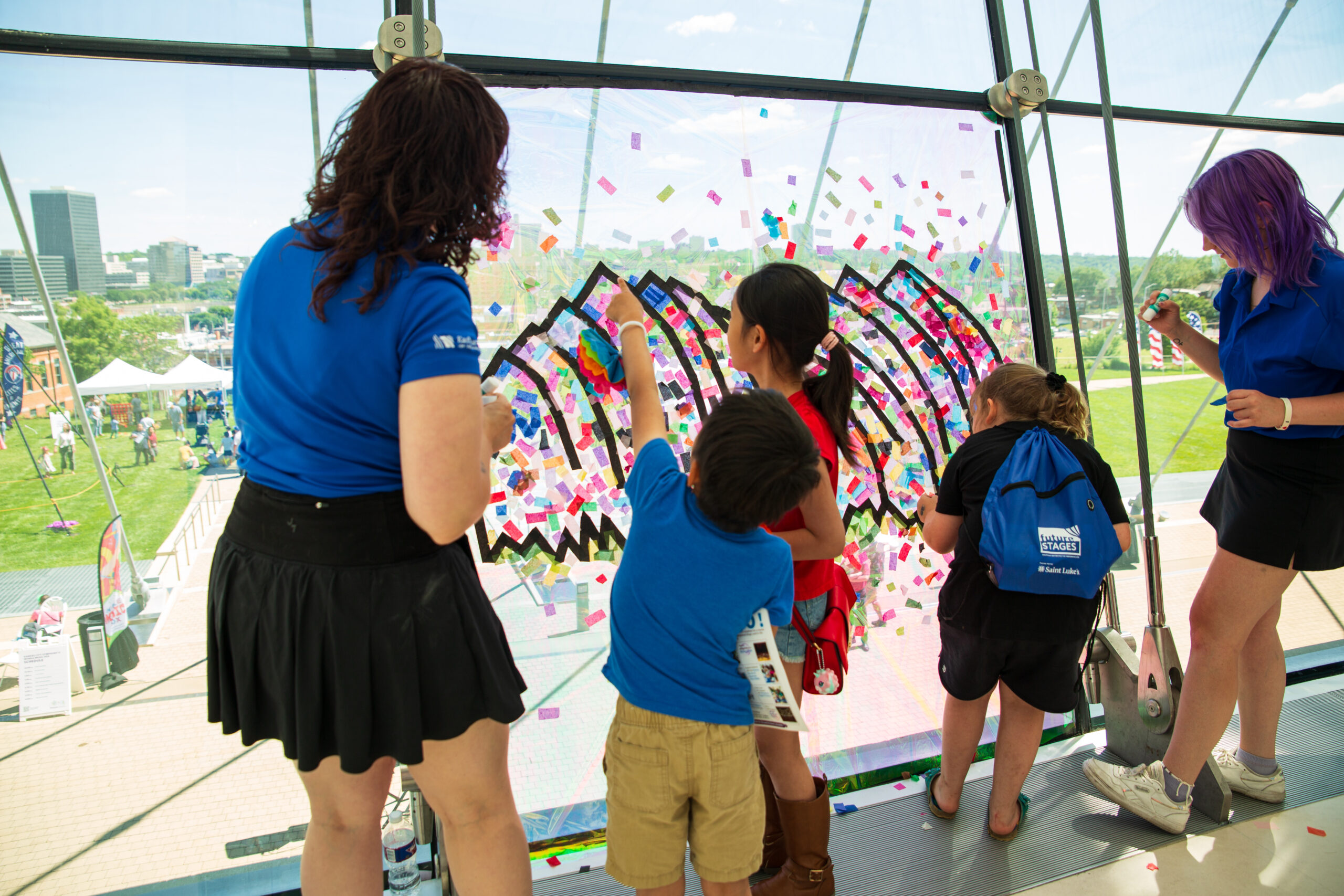 Children and adults participate in a collaborative art activity, using colorful sticky notes on a large transparent panel, during an outdoor event in a city park. Buildings and green space are visible outside.