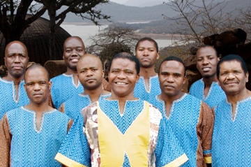 A group of nine men stand outdoors, wearing blue and brown traditional attire with a scenic landscape in the background.
