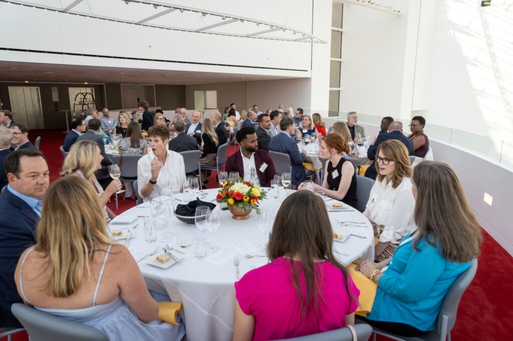 A large group of people are seated around multiple round tables covered with white tablecloths at a formal event. Each table has floral centerpieces and wine glasses. The attendees appear to be engaged in conversations, in a brightly lit area of Kauffman Center