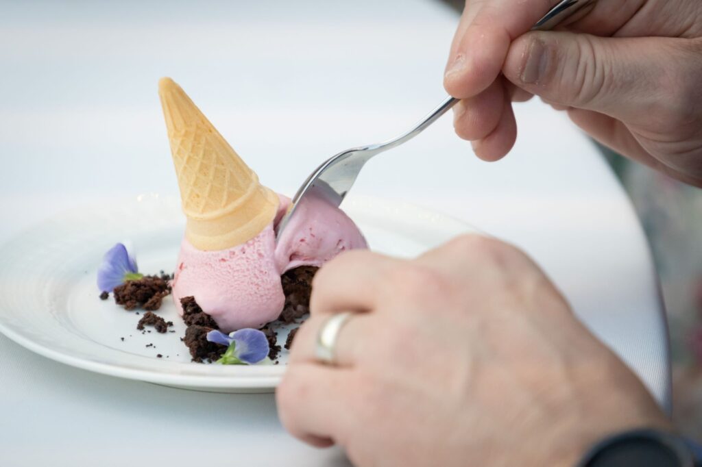A person is using a spoon to eat a creatively presented dessert resembling an upside-down ice cream cone with pink ice cream, chocolate crumbs, and edible flowers on a white plate. The person is wearing a ring on their left hand.