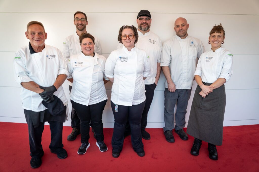 A group of seven chefs standing in a row against a white wall on a red carpet. They are all wearing white chef jackets, and some have additional accessories like glasses, hats, or aprons. They are smiling and looking at the camera.