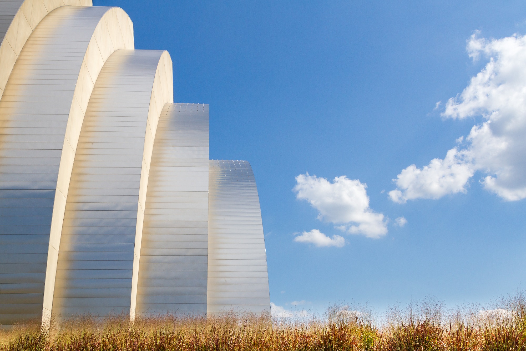 A modern building with curved, layered white walls under a blue sky with a few clouds, framed by grass in the foreground. Media credits capture the sleek design and natural surroundings perfectly.