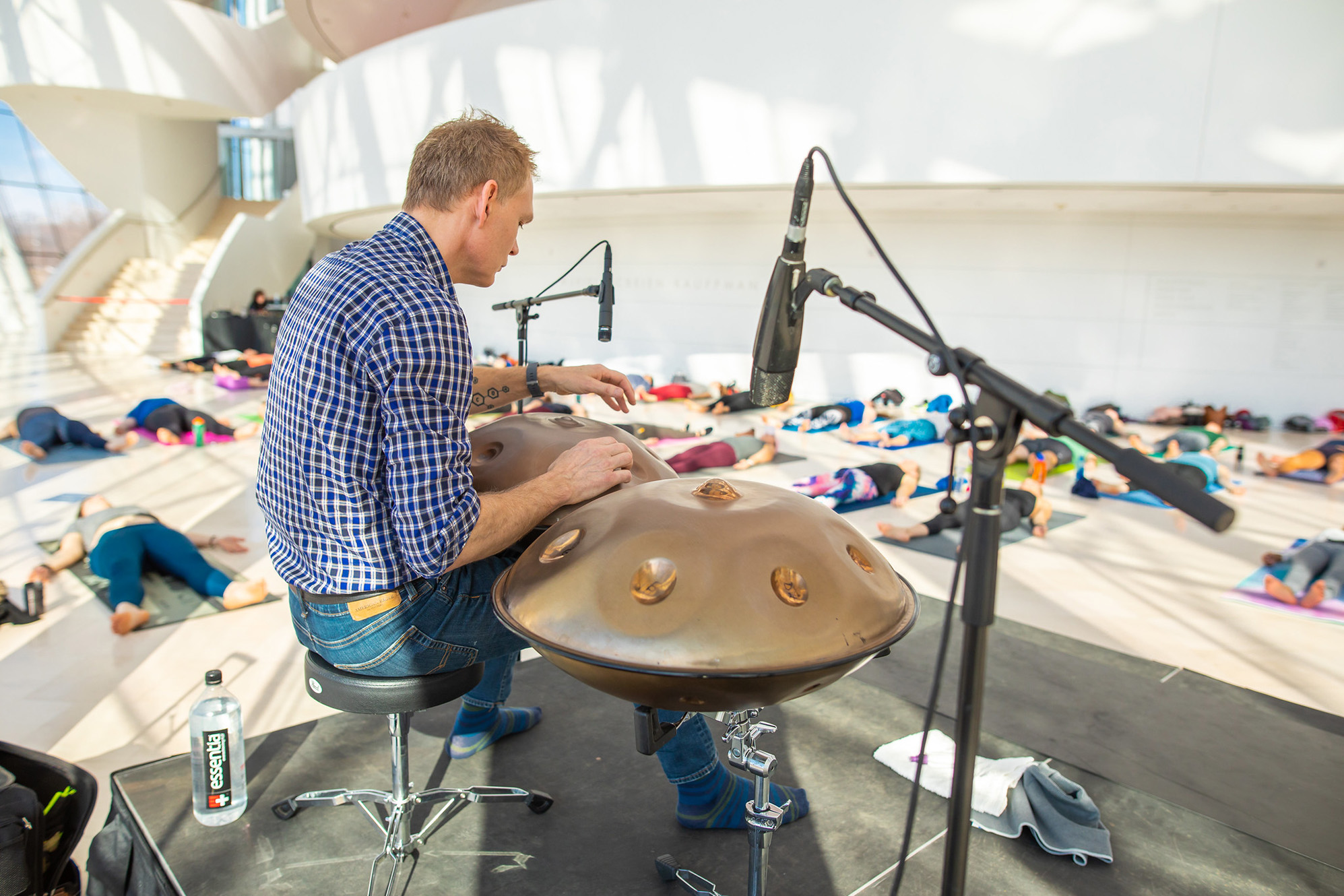 A person plays a handpan on a stage with a microphone while a group of people lie on yoga mats in a large, well-lit room during Yoga at Kauffman Center.