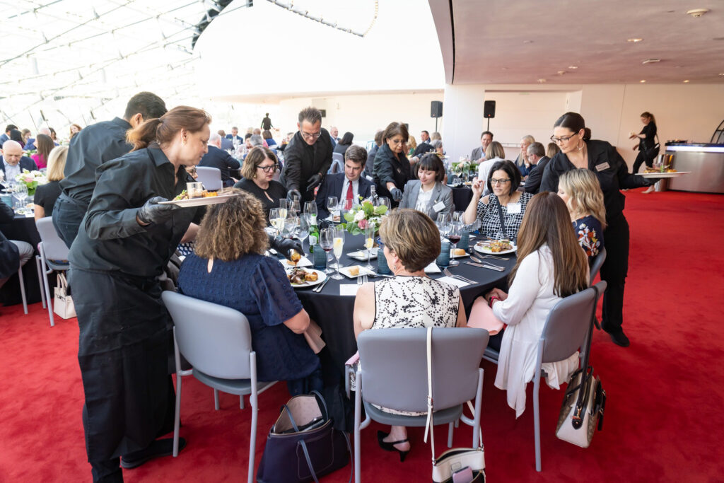 A group of people are seated around a table with white tablecloths at a formal event, likely discussing business memberships. Servers are delivering food and drinks to the table. The room has red carpeting and a modern design.