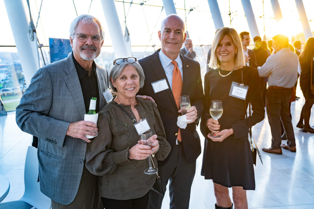 Four people, three holding wine glasses and one holding a beer, stand together smiling at an indoor business event with large windows and other attendees in the background, showcasing the camaraderie fostered through business memberships.
