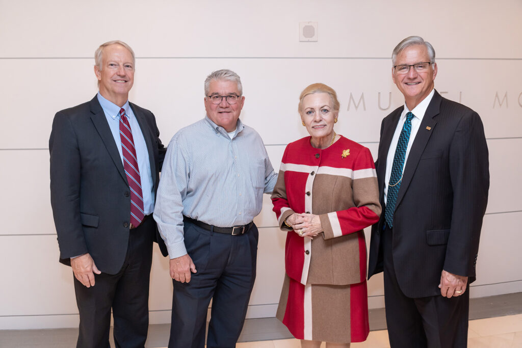 Four people stand side by side, three men in suits and one woman in a red and beige coat, all smiling at the camera. They are indoors against a plain background, likely celebrating their business memberships.
