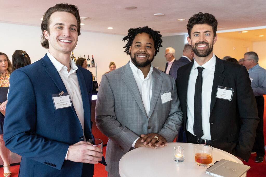Three men in suits at a networking event are standing around a small table, each holding a beverage. Two of them have name tags visible, indicating their business memberships. More attendees are seen in the background.