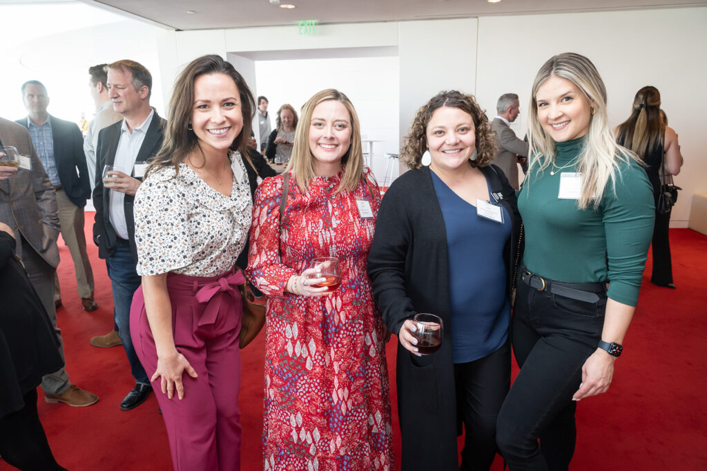 Four individuals are standing together on a red carpet, smiling, and holding drinks at a social event celebrating business memberships. Others are visible in the background.
