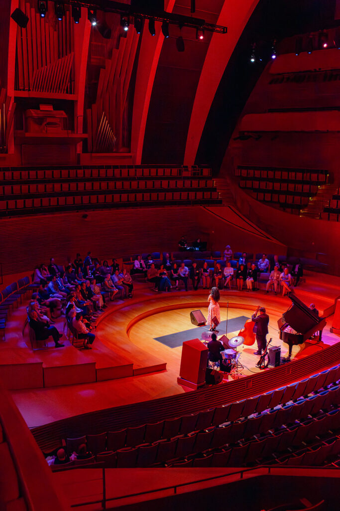 A jazz trio performs on a circular stage under red lighting in a modern auditorium with an audience, including some business memberships, seated around the stage.