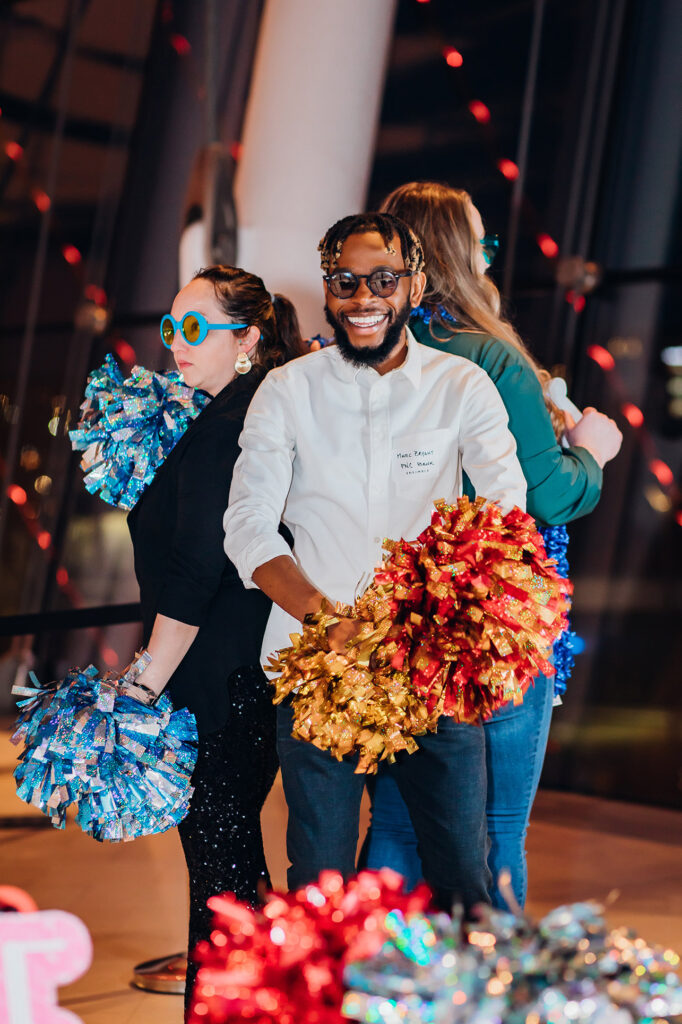 Three people are posing with colorful pom-poms indoors; the man in the foreground is smiling and wearing glasses, radiating a sense of camaraderie often seen in business memberships.