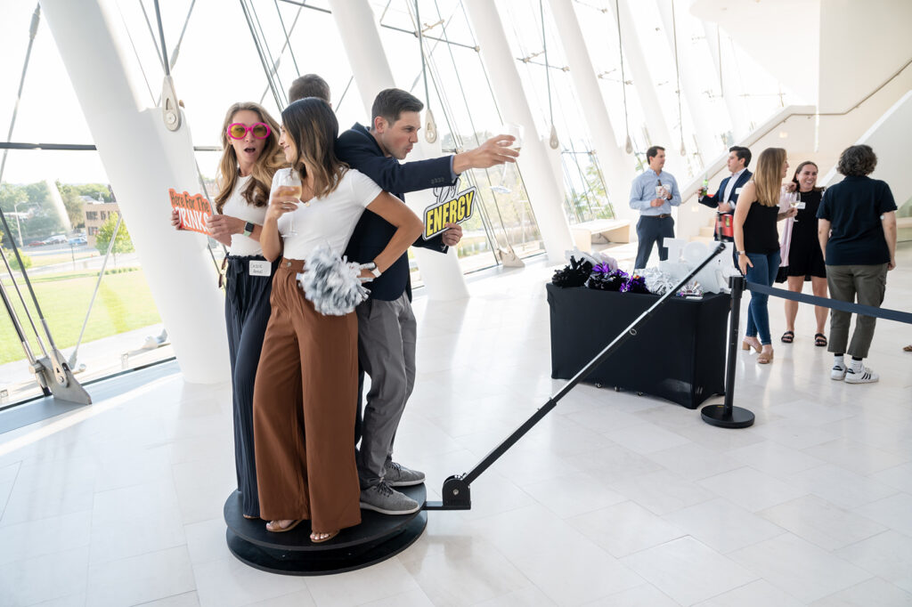 A group of three people poses with props in front of a photographer, while others mingle in the background of a brightly lit indoor event space, celebrating their business memberships.