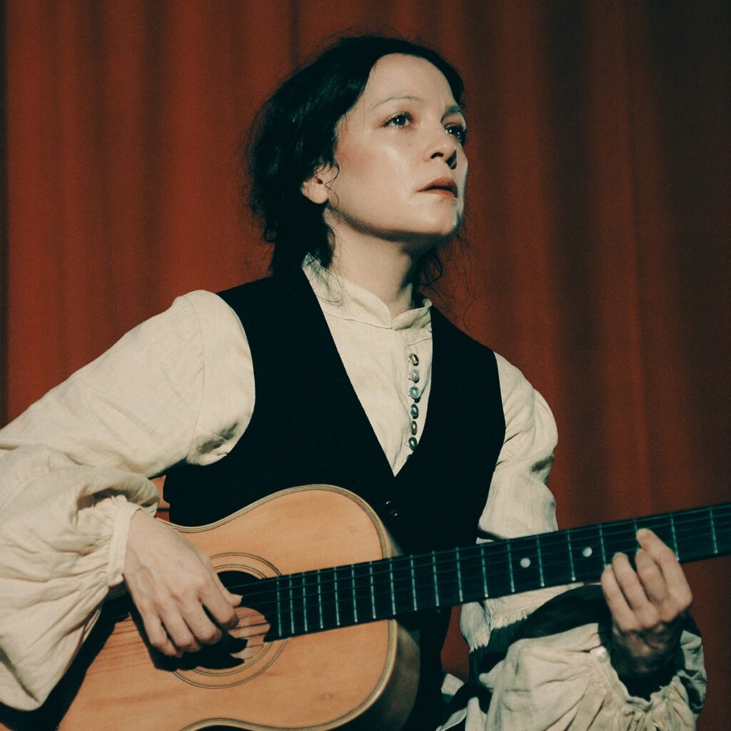 Natalia Lafourcade poses with an acoustic guitar with a red curtain in the background.