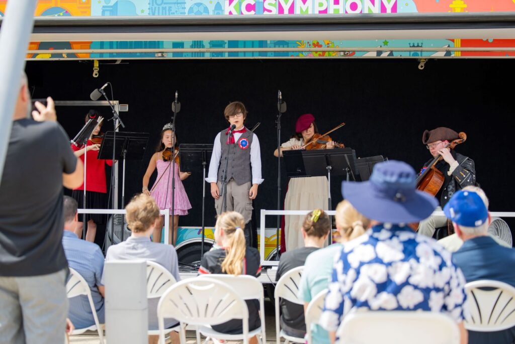 A group of musicians, including children and adults in costume, perform on stage outdoors for an audience seated in white chairs.