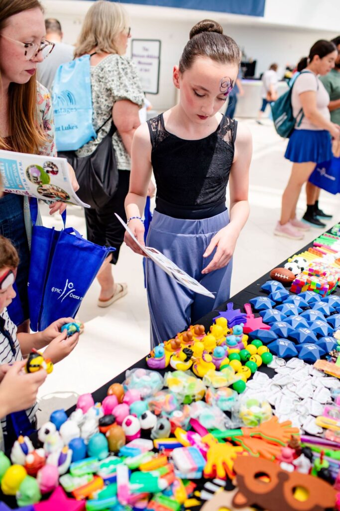 A girl stands at a table covered with colorful toys and trinkets, holding a paper and looking at the items, while other people browse in the background.