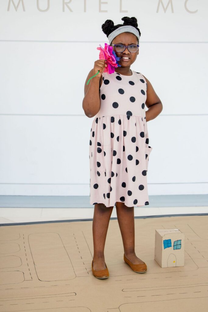 A young girl in a polka dot dress stands on a drawn street map, holding a pink flower and standing next to a small cardboard house.