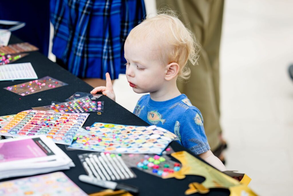 A young child in a blue shirt stands at a table covered with colorful sticker sheets and craft supplies.