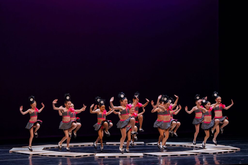 A group of tap dancers in matching costumes performs a synchronized tap dance routine on stage, each raising one knee and arms in similar poses.