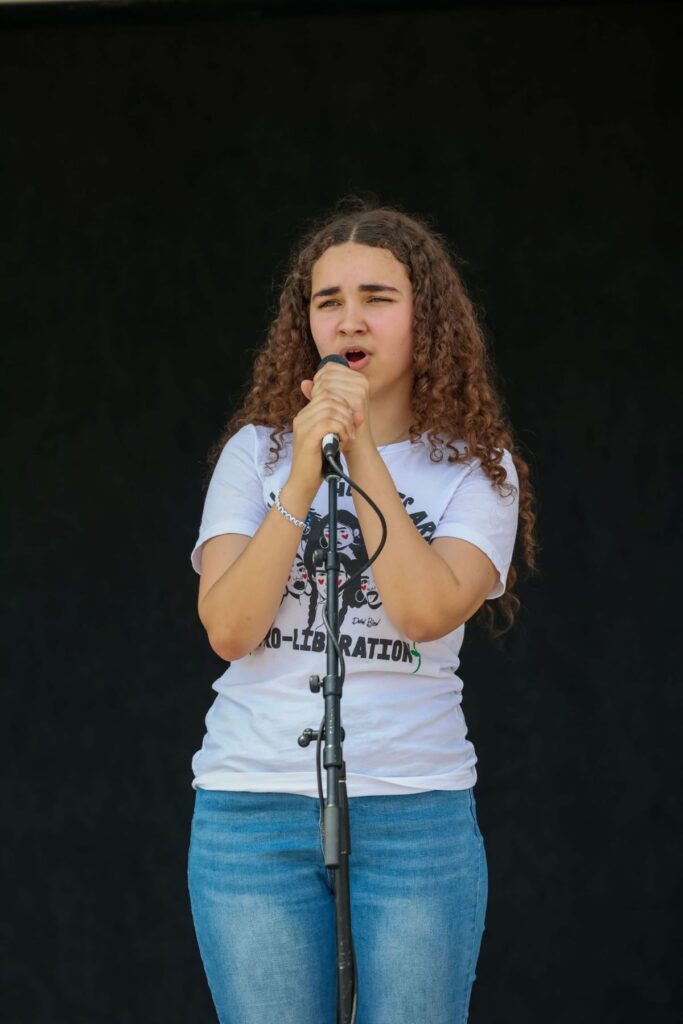 A young person with curly hair sings into a microphone on stage, wearing a white printed t-shirt and blue jeans against a plain dark background.