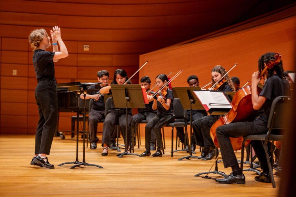 A conductor leads a small orchestra of young musicians playing string instruments on a stage in Helzberg Hall.