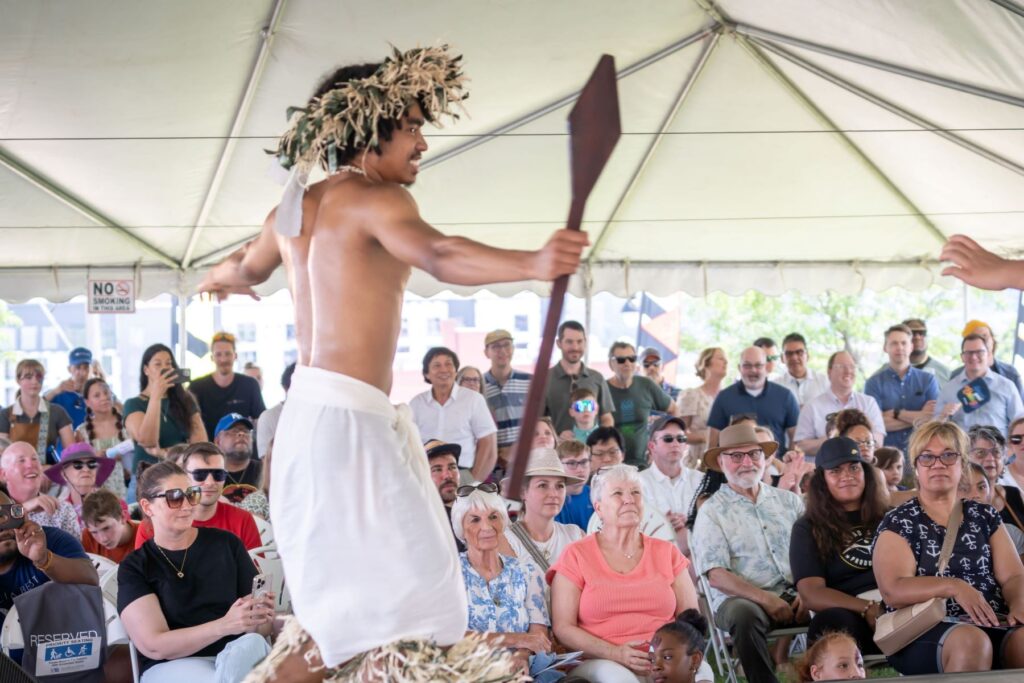 A performer dances on stage under a tent, while an audience watches attentively.