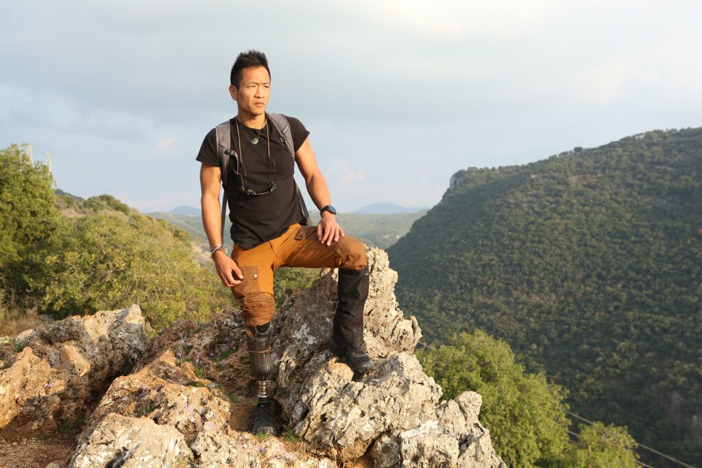 Albert Lin stands with a prosthetic leg stands on rocky terrain, wearing outdoor clothing and a backpack, with green hills in the background.
