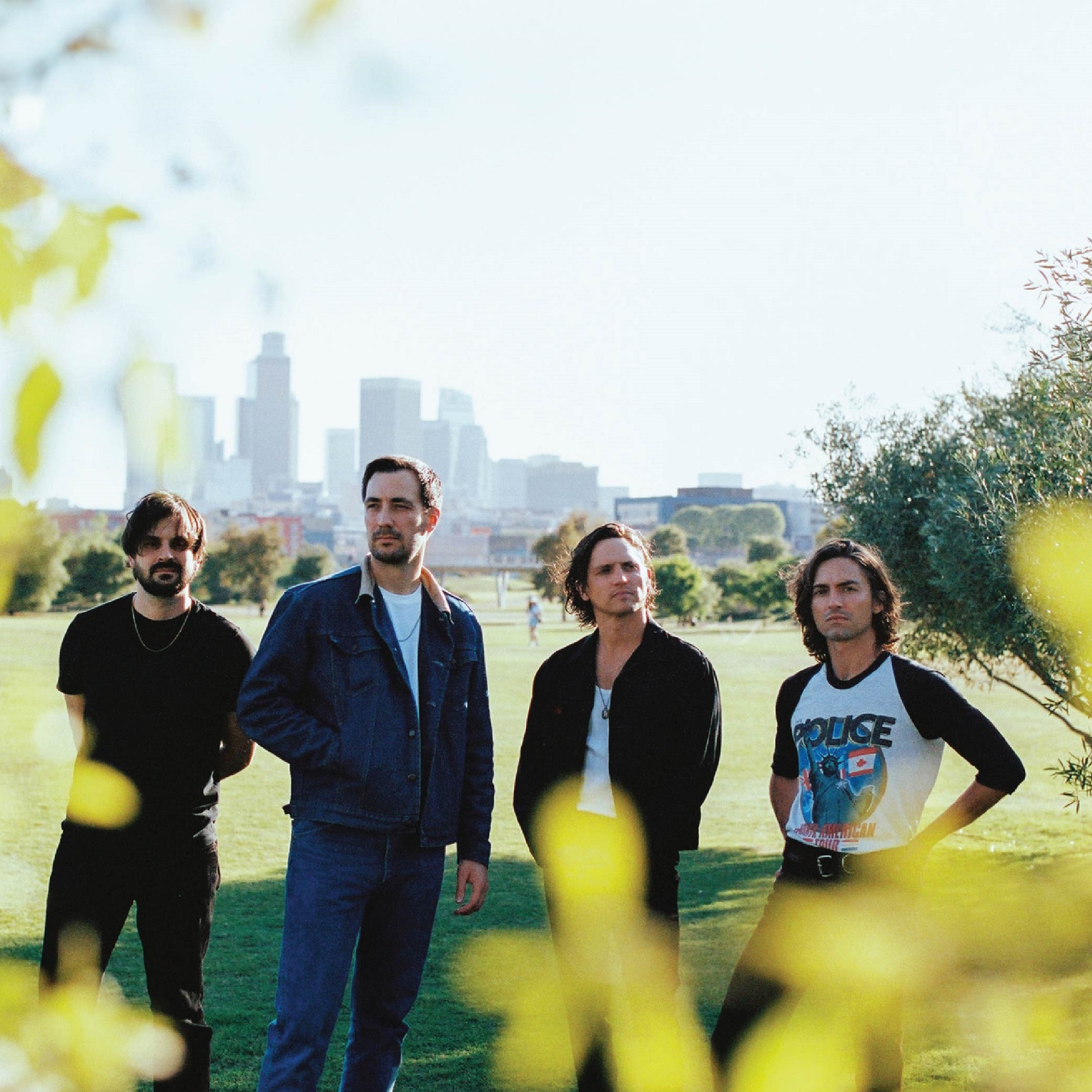 Four men stand in a grassy park with a city skyline in the background, framed by green foliage in the foreground.