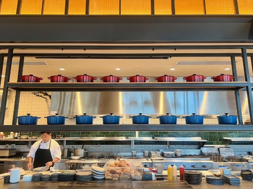 A restaurant kitchen with a chef working at the counter, and two rows of red and blue Dutch ovens displayed on a shelf above.