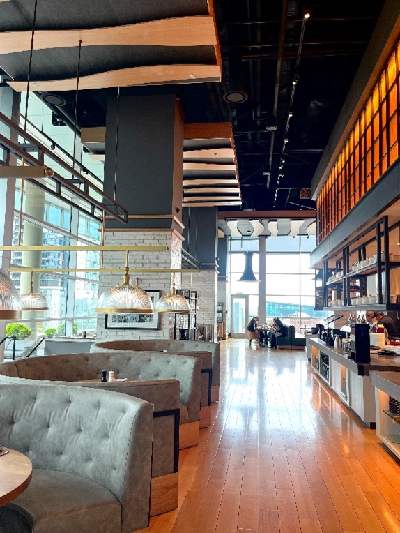 Modern restaurant interior with grey booths, wooden floors, large windows, and high ceilings with exposed beams; a few people are seated near the windows.