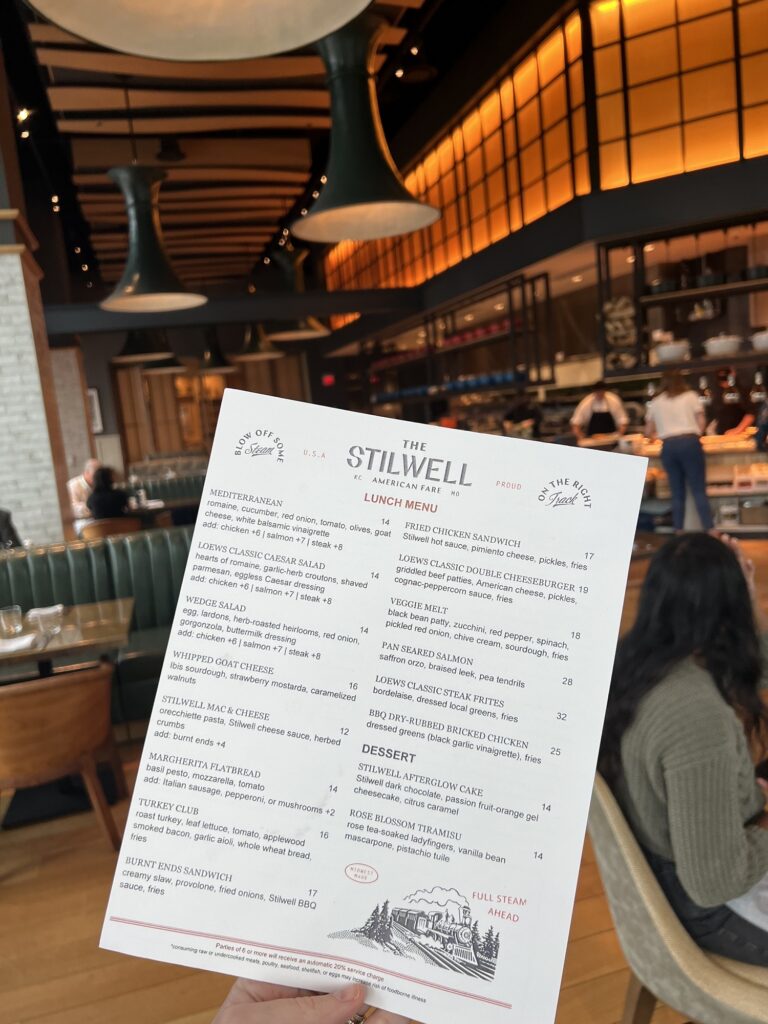A person holds up a lunch menu from The Stilwell restaurant inside a modern, warmly lit dining area with tables, chairs, and large overhead lights.