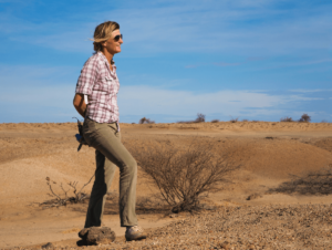 Louise Leakey wearing sunglasses and outdoor clothing stands in a dry, desert landscape with sparse vegetation under a blue sky.