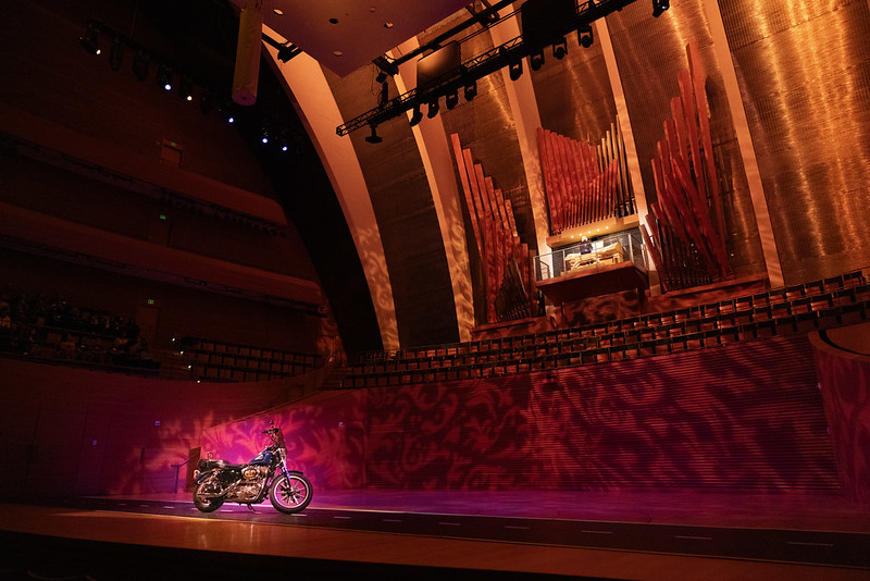 A motorcycle is displayed on stage under dramatic lighting at a PNC pop up event, set in front of a large pipe organ inside a modern concert hall.