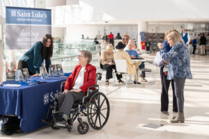 A woman in a wheelchair talks to a representative from Saint Luke’s Bishop Spencer Place at a booth in a bright lobby during Alzheimer's Awareness Month as others socialize in the background.