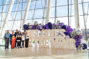 A group of people stands in front of a large “Melodies of the Mind” sign, decorated with purple and silver balloons, inside a glass-walled building for a free organ concert in honor of Alzheimer’s Awareness Month.