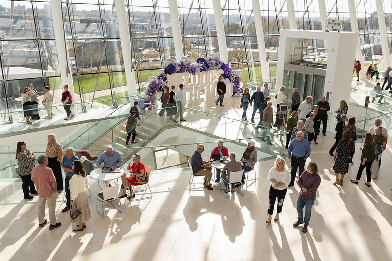 People gather and converse inside a spacious, modern building with large windows and a decorative purple balloon arch in the background.