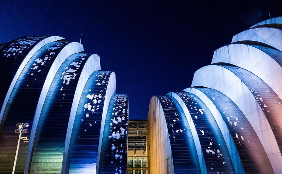 A modern building with large, curved metallic segments illuminated by blue lights at night, set against a dark sky — the Kauffman Center stands out beautifully.