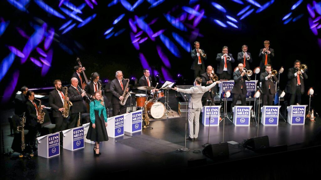 A large jazz band performs on stage, with musicians playing brass, woodwind and percussion instruments. The conductor stands in front and two vocalists are positioned near the center.
