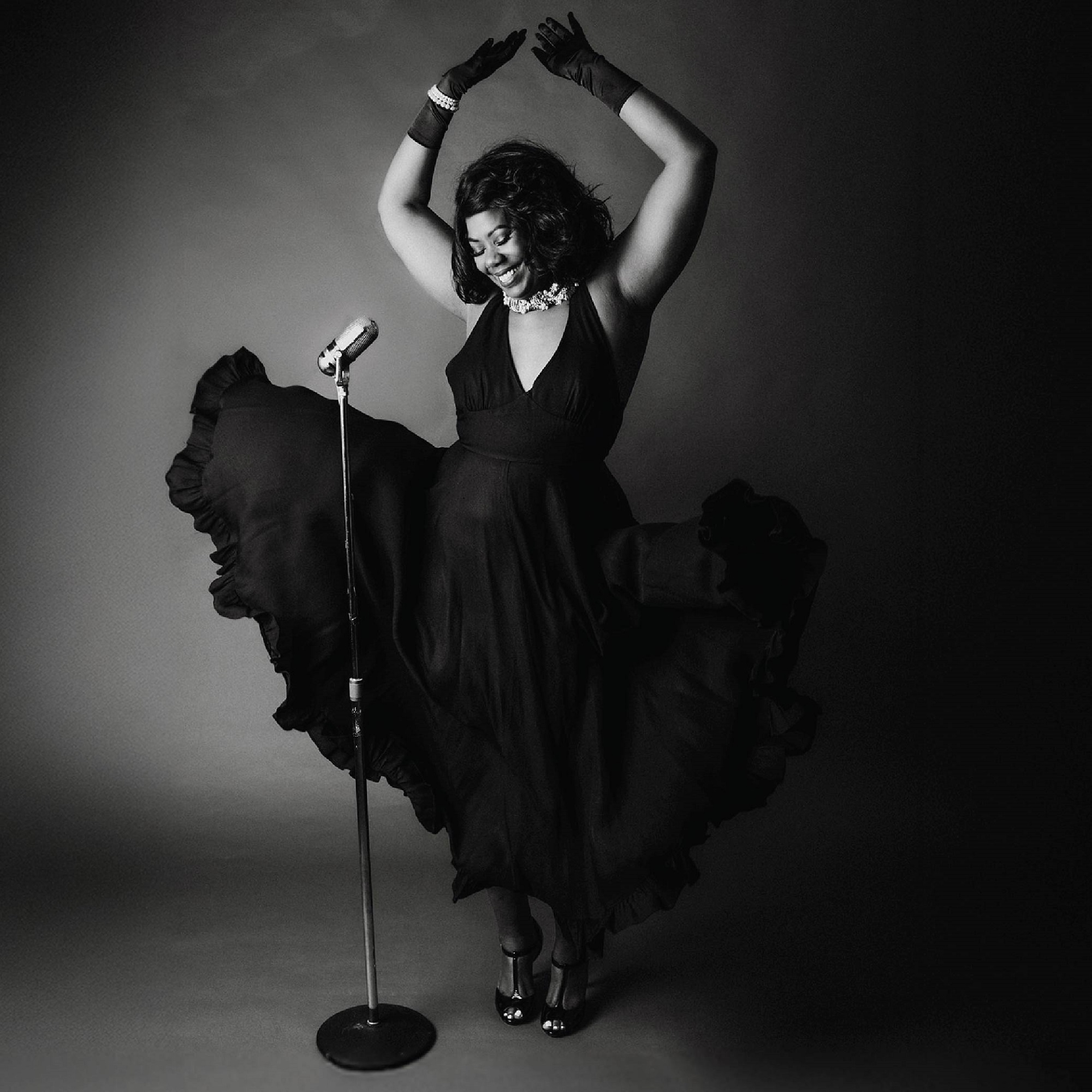 A woman in a flowing black dress poses with arms raised, standing in front of a vintage microphone on a stand in a studio setting.