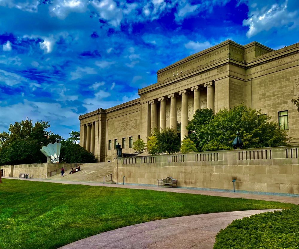 A large stone building with columns, surrounded by trees and grass, under a partly cloudy blue sky. People are visible on the steps and benches near the entrance.