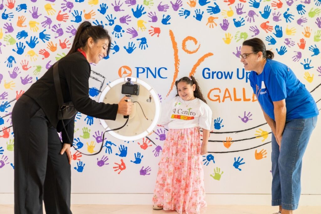 Three people pose for a photo in front of a colorful handprint wall; one woman holds a phone with a ring light, while the girl and another woman smile.