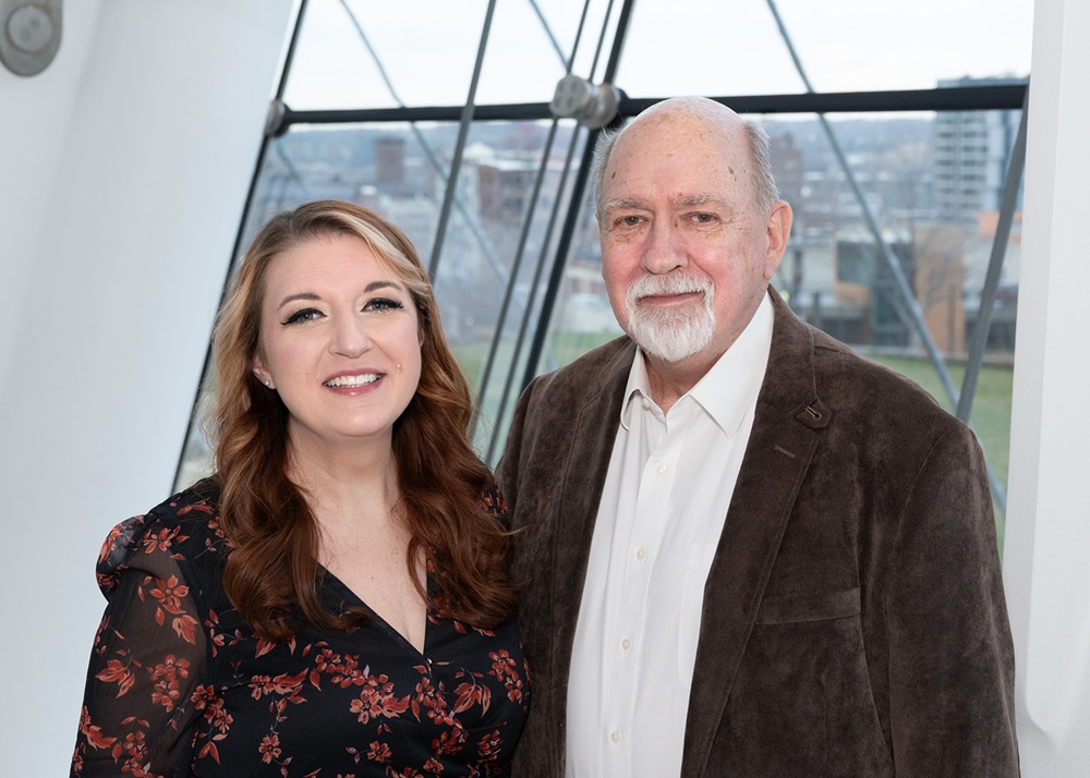 A woman and an older man stand side by side indoors in front of large windows with a cityscape view in the background.
