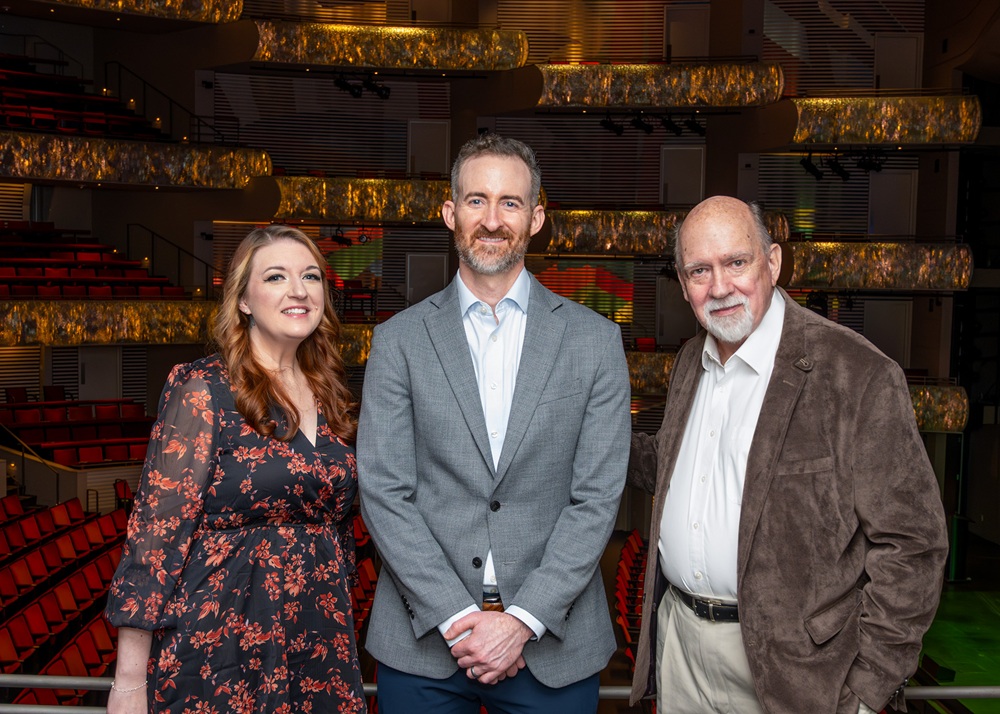 Three people stand side by side in a theater auditorium with red seats, posing for a photo. The background shows multiple rows of empty seats and balconies.