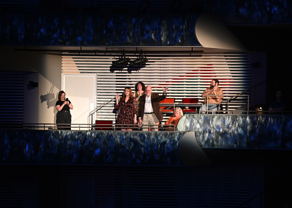 A group of people stands and sits on a balcony in a theater. One man is pointing, while others watch. A woman on the left signs with her hands.