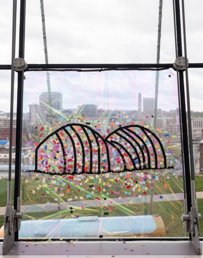 View through a window decorated with black arched lines and colorful confetti shapes, showing a cityscape with buildings under a cloudy sky in the background.