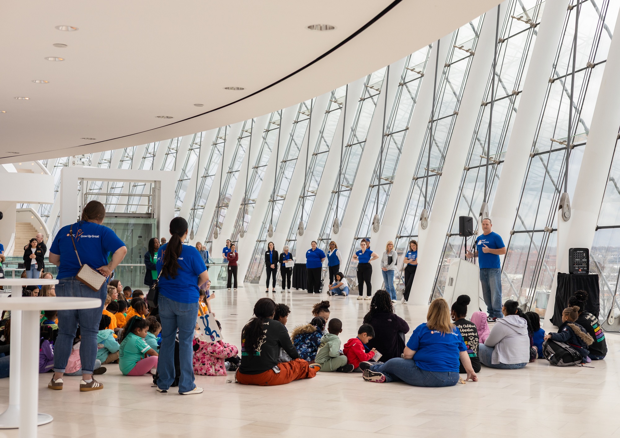 A group of adults in blue shirts stand and speak to seated children and adults in a large, modern, glass-walled atrium.