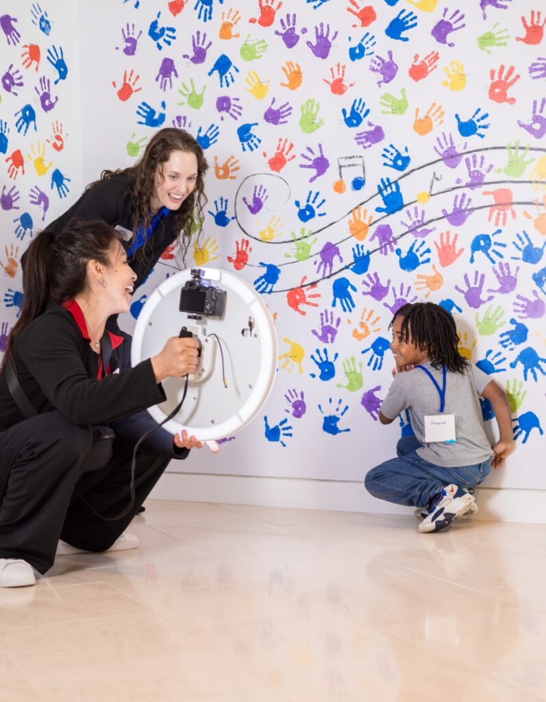 Two adults with a ring light and camera interact with a child in front of a wall covered in colorful handprints and a looping black line.