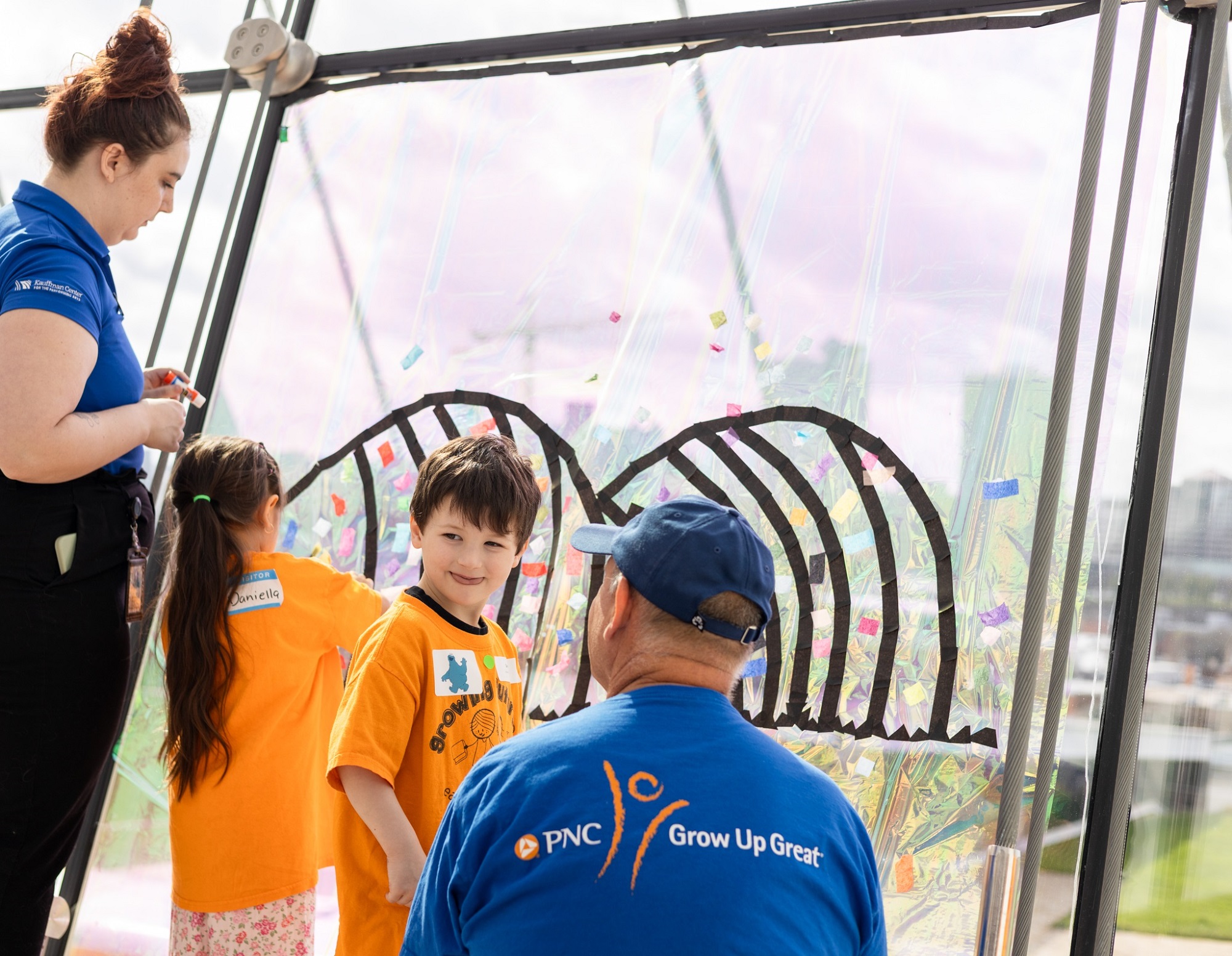 Two adults and two children work together to decorate a large, clear panel with colorful stickers and a black outlined design at an indoor event.