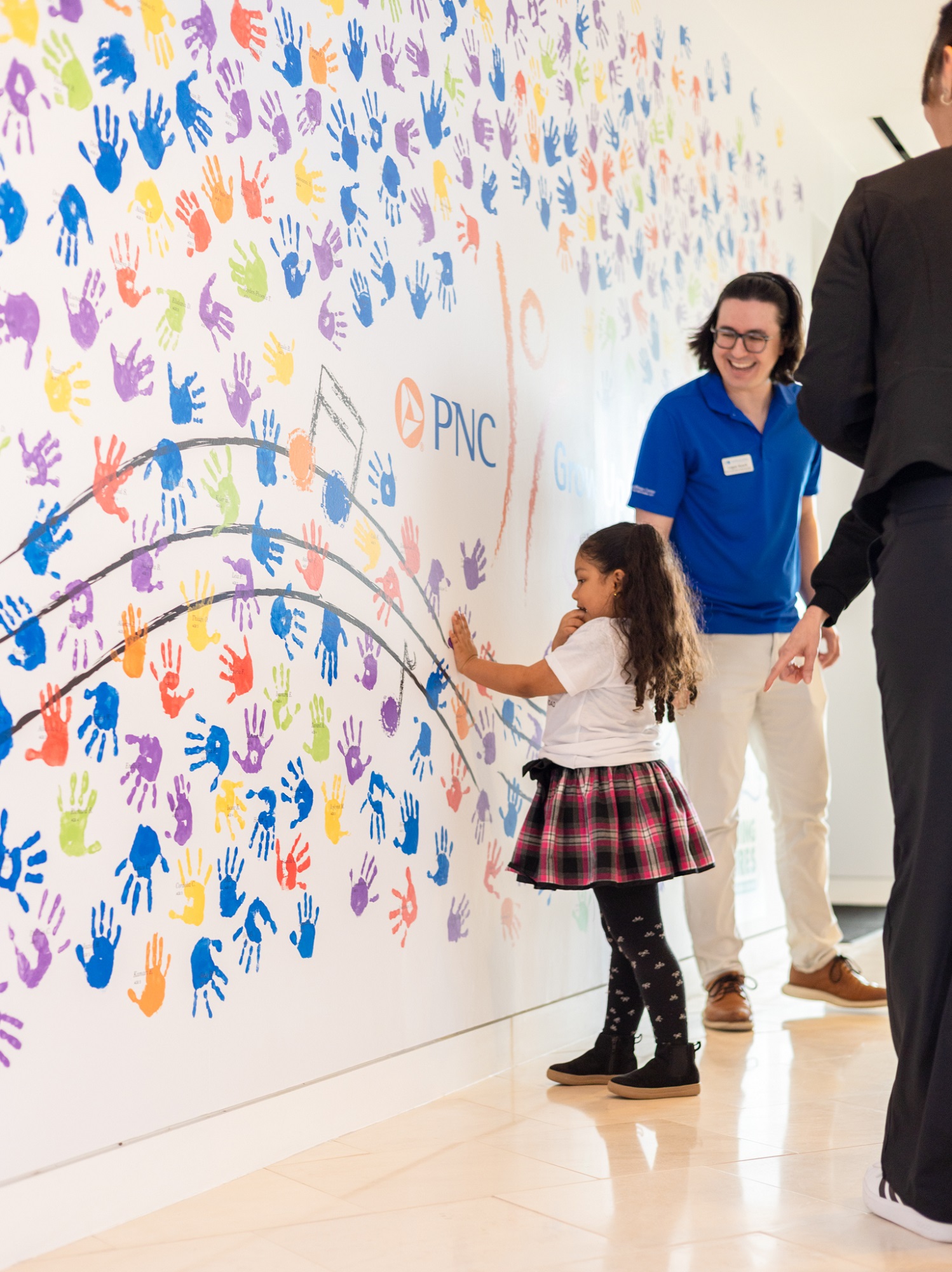 A young girl touches a wall covered in colorful handprints as adults supervise in a bright indoor space.