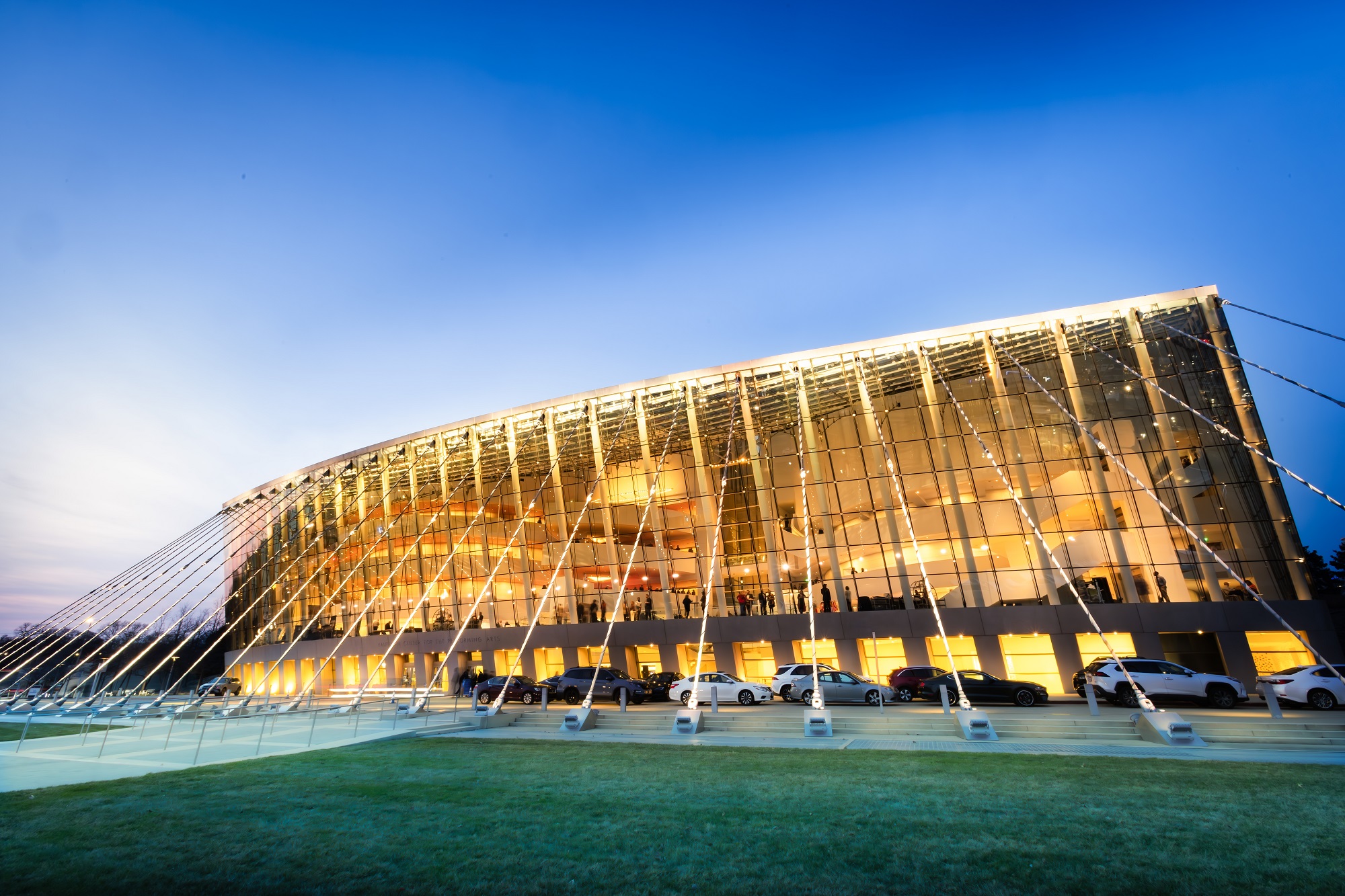 A modern glass-fronted building with large windows is illuminated at dusk, with a grassy area in the foreground.